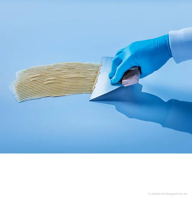 Eine behandschuhte Hand zeigt den Klebstoffauftrag mit einem Spachtel.