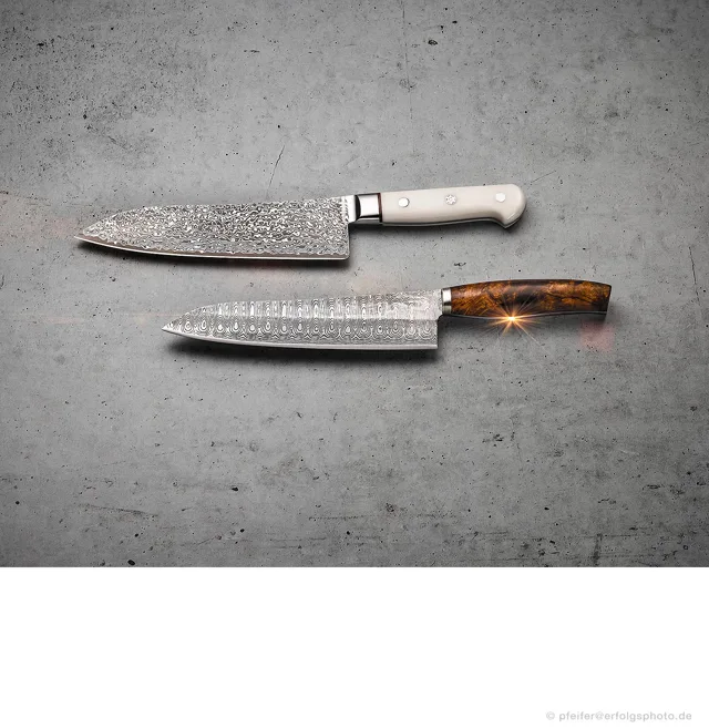 Zwei Küchenmesser zeigen ihre gemusterten Klingen vor einer strukturierten Fläche.